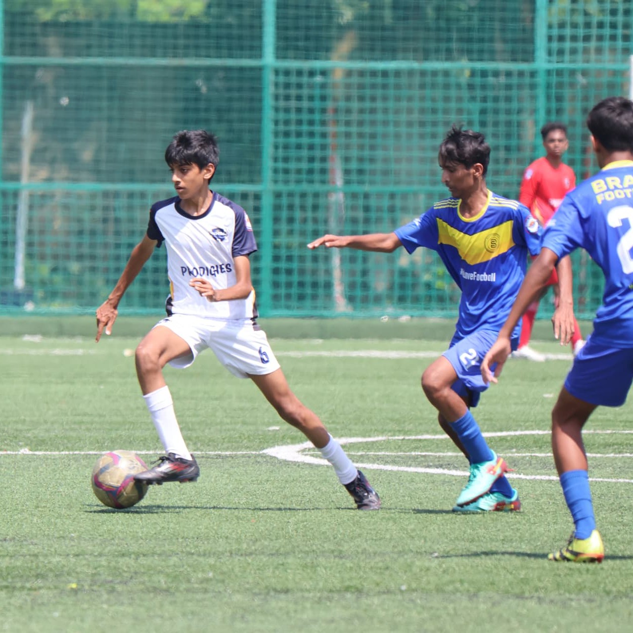 Apoorv Gupta dribbling the ball in a white Prodigies kit while a defender in blue closes in during a youth soccer match on turf